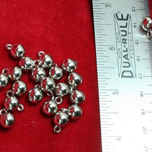 May include: A collection of small, shiny silver beads with loops, scattered on a red surface. A ruler is placed next to the beads for scale. The beads appear to be approximately 0.6 cm in diameter.