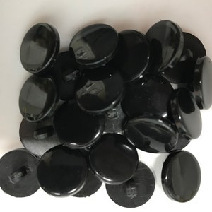 May include: A collection of glossy, black, round buttons. Each button has a flat, smooth surface and a small loop on the back for sewing. The buttons are scattered on a white surface.