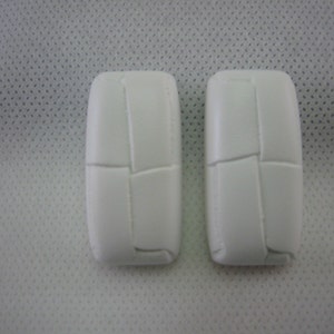 May include: Two white, rectangular objects with a textured, stitched design. The objects are positioned side-by-side against a white, dotted background. The objects have rounded edges and a segmented appearance.