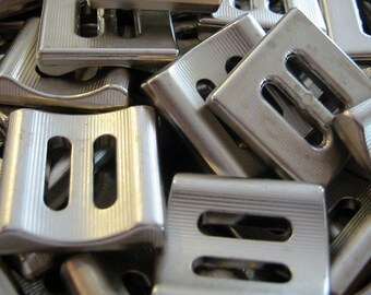 May include: Close-up of a pile of shiny, silver-colored metal buckles. Each buckle has a rectangular shape with two rectangular cutouts and a curved top. The buckles are stacked together, creating a textured surface.