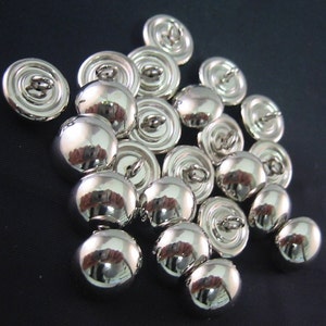 May include: A collection of shiny, silver-colored buttons with a domed front and a loop on the back for sewing. The buttons are reflective, with a smooth, polished surface. The buttons are scattered on a dark background.