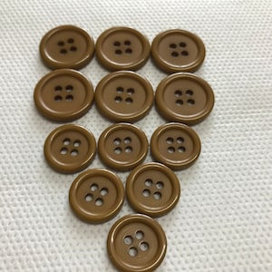 May include: A collection of eleven brown, round buttons with four holes each. The buttons are arranged in a slightly staggered pattern against a white, textured background. The buttons are a solid, matte color.