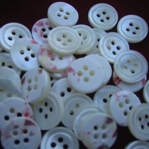 May include: A collection of off-white, four-hole buttons. Some buttons have a subtle pink and red pattern. The buttons vary in size and are scattered across a dark brown surface. These buttons are suitable for sewing and craft projects.