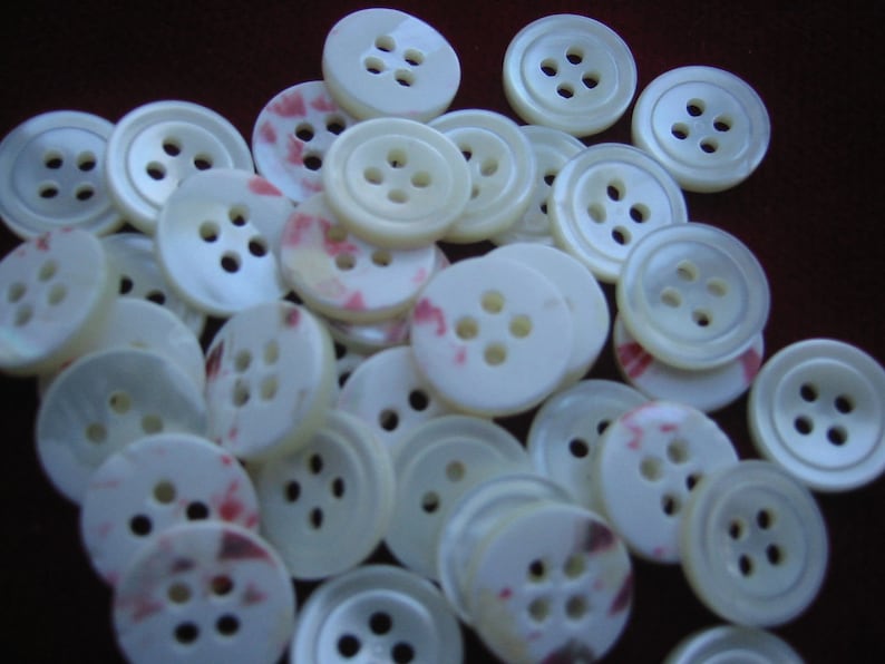 May include: A collection of off-white, four-hole buttons. Some buttons have a subtle pink and red pattern. The buttons vary in size and are scattered across a dark brown surface. These buttons are suitable for sewing and craft projects.
