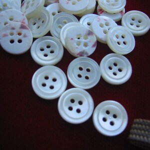 May include: A collection of white buttons with four holes each. Some buttons have a subtle pink and purple pattern. The buttons are scattered on a dark red surface, suggesting a craft or sewing project.