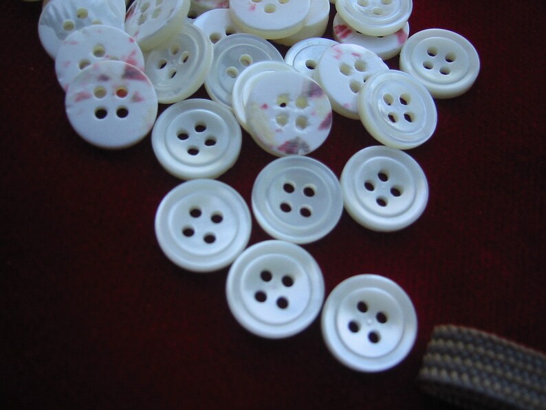 May include: A collection of white buttons with four holes each. Some buttons have a subtle pink and purple pattern. The buttons are scattered on a dark red surface, suggesting a craft or sewing project.