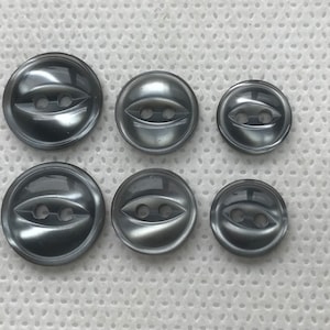 May include: Six gray buttons of varying sizes are arranged in two rows. Each button has a rounded shape with a central, eye-shaped design and two holes for sewing. The buttons have a glossy finish.