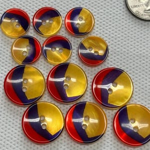 May include: A collection of colorful buttons with a unique design. Each button is round, featuring a split design with gold, red, and purple sections. The buttons have two holes for sewing and a glossy finish. A quarter is in the background.