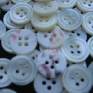 May include: A collection of white buttons with four holes each. Some buttons have a slight pearlescent sheen, while others have a subtle pink and purple pattern. The buttons are scattered, creating a textured surface.