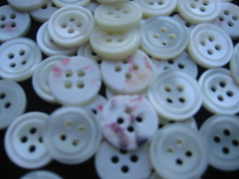 May include: A collection of white buttons with four holes each. Some buttons have a slight pearlescent sheen, while others have a subtle pink and purple pattern. The buttons are scattered, creating a textured surface.