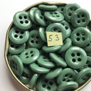May include: A collection of sage green buttons with four holes each, displayed in a small, round, metal container. A small, yellow sticky note with the number "53" is visible. These buttons are suitable for sewing and craft projects.