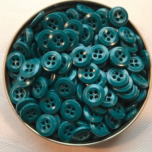 May include: A collection of teal-colored, four-hole buttons fills a small, round metal container. The buttons are evenly sized and have a glossy finish. The container is slightly tilted, revealing the buttons' arrangement.