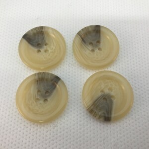 May include: Four round buttons with a marbled design in shades of beige and brown. Each button has four holes for sewing. The buttons are arranged in a square formation.
