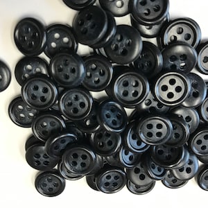 May include: A collection of black, round buttons with four holes each. The buttons are made of a smooth, glossy material and are scattered across a white surface. These buttons are suitable for sewing and craft projects.