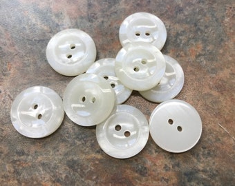 May include: Ten off-white, round buttons with two holes each. The buttons have a pearlescent sheen and are scattered on a textured, brown surface. These buttons are suitable for sewing and craft projects.