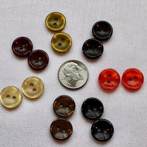 May include: A collection of round buttons in various colors, including amber, brown, black, and orange. Each button has two holes for sewing. A U.S. dime is included for size comparison. The buttons are made of a shiny, smooth material.
