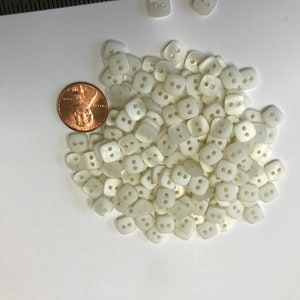 May include: A collection of small, square, off-white buttons with two holes each, scattered on a white surface. A U.S. penny is included for size comparison. These buttons are likely for sewing or crafting projects.