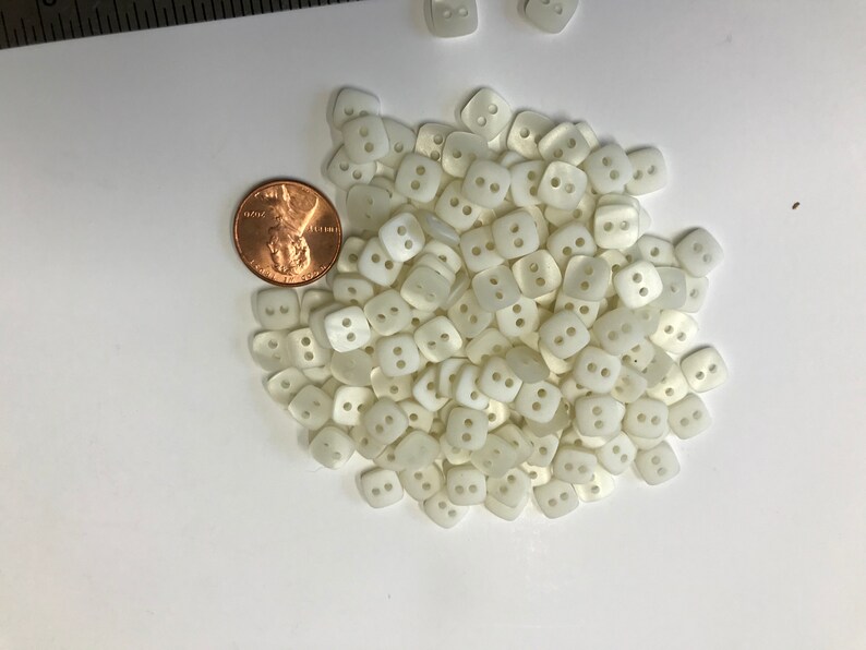 May include: A collection of small, square, off-white buttons with two holes each, scattered on a white surface. A U.S. penny is included for size comparison. These buttons are likely for sewing or crafting projects.