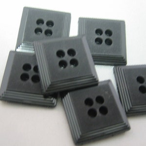 May include: A collection of six square, black buttons. Each button has four holes and a stepped, ridged border. The buttons are arranged in a slightly overlapping pattern, showcasing their design and texture. Ideal for sewing and craft projects.