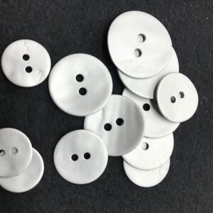 May include: A collection of various sized, round, light gray buttons with two holes each. The buttons are scattered on a dark gray surface. The buttons are made of a smooth, matte material.