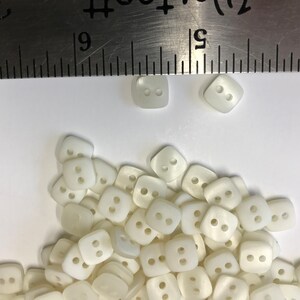 May include: A collection of off-white, square buttons with two holes each. A metal ruler is placed above the buttons, showing measurements in inches. The buttons are scattered on a white surface, suitable for sewing and craft projects.