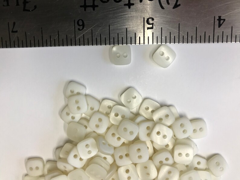 May include: A collection of off-white, square buttons with two holes each. A metal ruler is placed above the buttons, showing measurements in inches. The buttons are scattered on a white surface, suitable for sewing and craft projects.