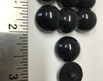 May include: A collection of glossy black buttons, some arranged in a cluster. The buttons are round with a slightly domed surface. A ruler is placed next to the buttons for scale. The buttons are approximately 1 inch in diameter.