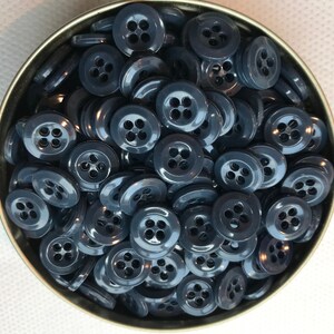 May include: A container filled with numerous navy blue buttons. Each button is round with four holes and a slightly raised edge. The buttons are made of a smooth, glossy material, and are scattered throughout the container.