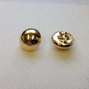 以下が含まれることがあります： 光沢のある金色のボタンが2つ。1つのボタンは滑らかで丸みを帯びており、光を反射しています。もう1つのボタンには、縫い付け用のループと穴があります。これらのボタンは、おそらく衣類や工芸品用です。