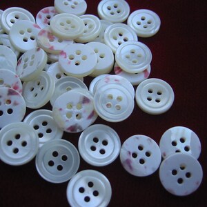 May include: A collection of off-white buttons with four holes each. Some buttons have a subtle pink and purple mottled pattern. The buttons are round and have a slightly pearlescent sheen, scattered on a dark red-brown surface.