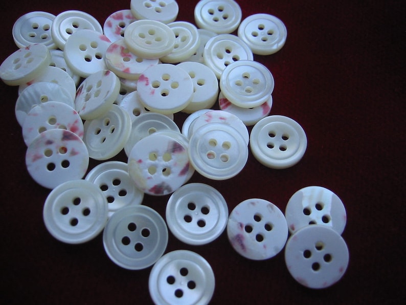 May include: A collection of off-white buttons with four holes each. Some buttons have a subtle pink and purple mottled pattern. The buttons are round and have a slightly pearlescent sheen, scattered on a dark red-brown surface.