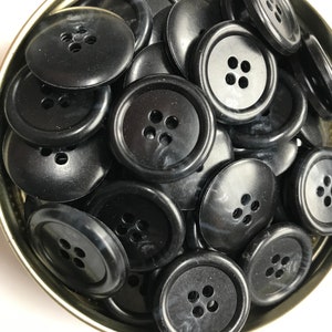 May include: A collection of black, round buttons with four holes each, inside a silver-colored tin. The buttons have a slightly glossy finish and are densely packed together. Ideal for sewing projects or crafts.