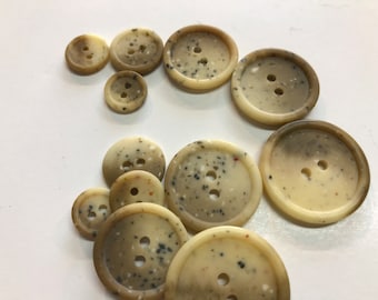 Puede incluir: Una colección de botones de dos agujeros de varios tamaños. Los botones son de color beige moteado con motas marrones oscuras y negras. Están dispuestos sobre una superficie blanca.
