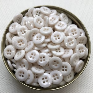 May include: A collection of small, round, off-white buttons with four holes each, filling a small, round, metal container. The buttons have a glossy finish and are scattered throughout the container, creating a textured appearance.