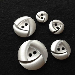 May include: A collection of five silver-colored buttons with a unique, geometric design. Each button features two holes and a swirling, triangular pattern. The buttons are arranged on a black surface.