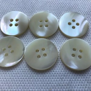 May include: Six round, off-white buttons with four holes each. The buttons have a pearlescent sheen and are arranged on a white, textured surface. These buttons are suitable for sewing and craft projects.