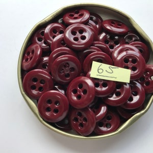 May include: A collection of burgundy-colored buttons with four holes, displayed in a tarnished metal container. A small yellow tag with the number "65" is visible. These buttons are suitable for sewing and craft projects.