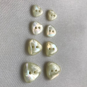 May include: A collection of eight pearlescent, triangular buttons with two holes each. The buttons have a subtle iridescent sheen, reflecting light with hints of pink, green, and blue. They are arranged on a white, textured surface.