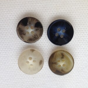May include: Four round buttons in various colors: dark brown, navy blue, off-white, and tan. Each button has four holes for sewing. The buttons have a marbled appearance, adding a touch of visual interest to the design.