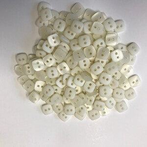 May include: A pile of off-white, square buttons with two holes each. The buttons are slightly rounded and have a matte finish. They are scattered across a white surface, suggesting a craft or sewing project.