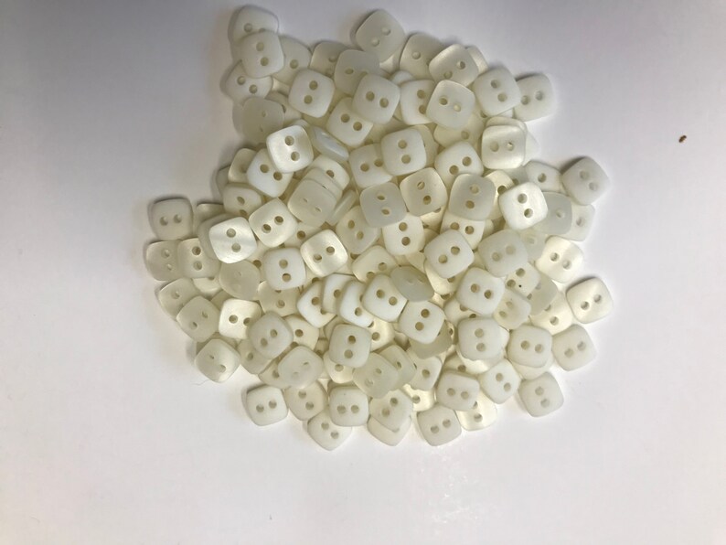 May include: A pile of off-white, square buttons with two holes each. The buttons are slightly rounded and have a matte finish. They are scattered across a white surface, suggesting a craft or sewing project.