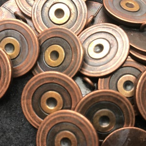 Op de afbeelding: Een verzameling metalen knopen in antieke stijl. Elke knoop is cirkelvormig met concentrische ringen van koper, donker brons en een messing midden. Sommige knopen hebben een lus aan de achterkant om te naaien.