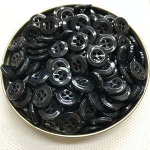 May include: A collection of glossy black buttons with four holes each, filling a small, round, metal container. The buttons are evenly sized and have a slightly raised edge.
