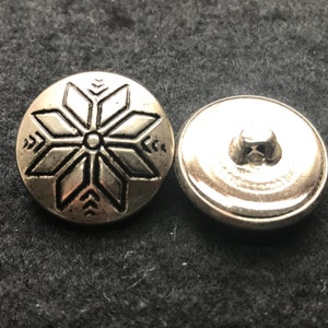May include: Two silver-coloured metal snap buttons. Each button's front has a raised, stylised snowflake design. The buttons are round and have a slightly aged appearance. The background is a dark, textured surface.