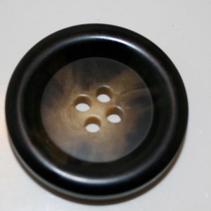 May include: A large, round button with four holes. The button has a dark brown outer rim and a lighter, mottled brown center. The button is likely made of plastic or a similar material.