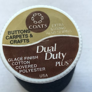 Puede incluir: Un carrete de hilo negro Dual Duty Plus. La etiqueta es blanca, marrón y dorada, con el texto "Buttons, Carpets & Crafts" y "Extra Strong Hand Sewing". El hilo está hecho en los EE. UU.