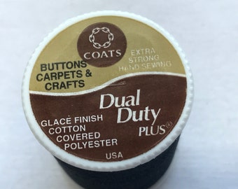 Puede incluir: Un carrete de hilo negro Dual Duty Plus. La etiqueta es blanca, marrón y dorada, con el texto "Buttons, Carpets & Crafts" y "Extra Strong Hand Sewing". El hilo está hecho en los EE. UU.