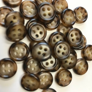 May include: A collection of round buttons with four holes each. The buttons have a mottled brown and tan pattern, with a lighter center. The buttons are scattered on a white surface.