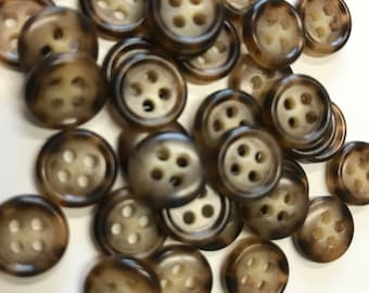 May include: A collection of round buttons with four holes each. The buttons have a mottled brown and tan pattern, with a lighter center. The buttons are scattered on a white surface.