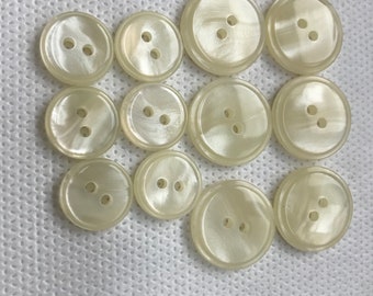 May include: Twelve off-white, pearlescent buttons with two holes each, arranged on a white, textured surface. The buttons vary in size and have a subtle, iridescent sheen. Ideal for sewing projects.